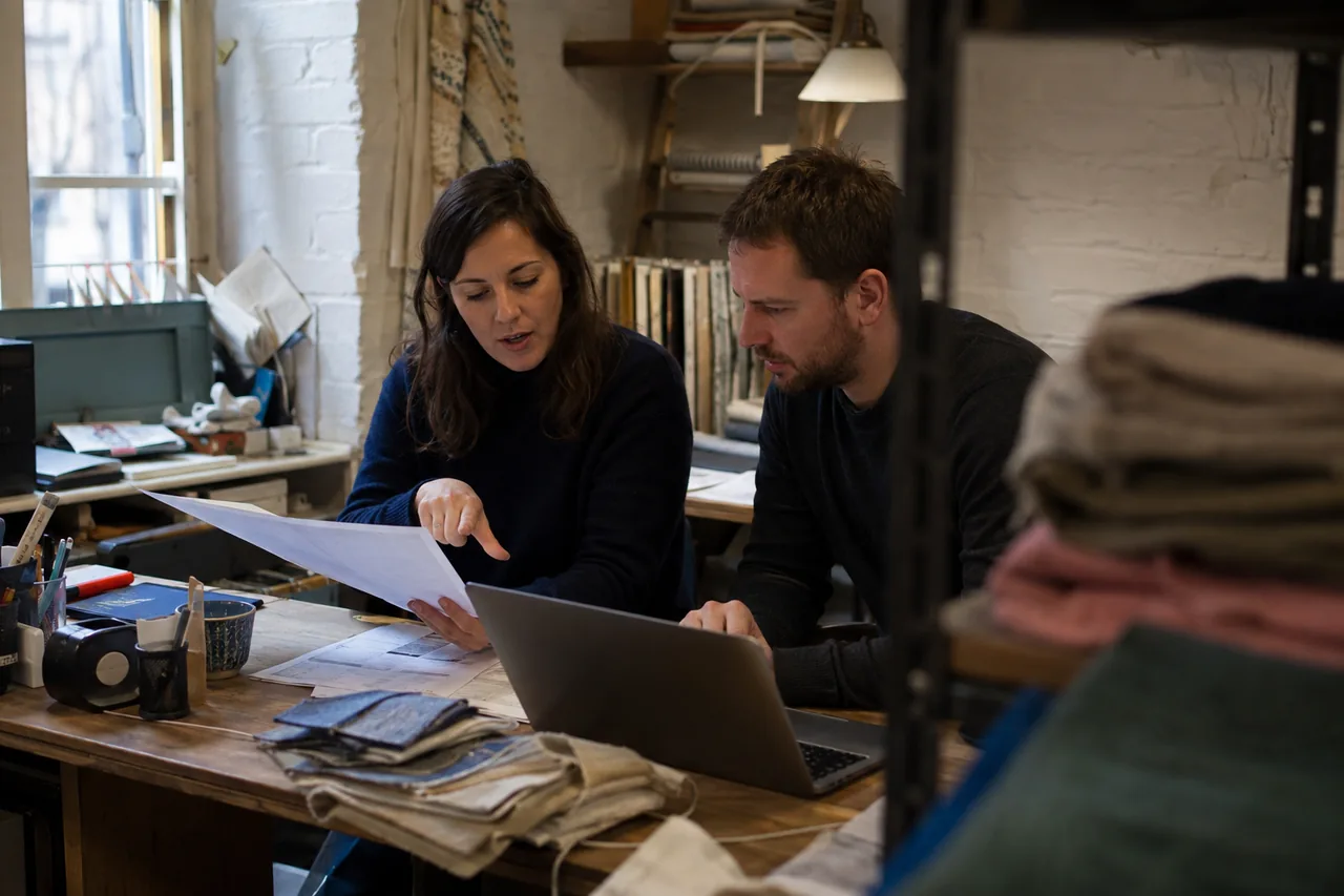 Two small-business colleagues reviewing a supplier document and laptop in a working textile studio